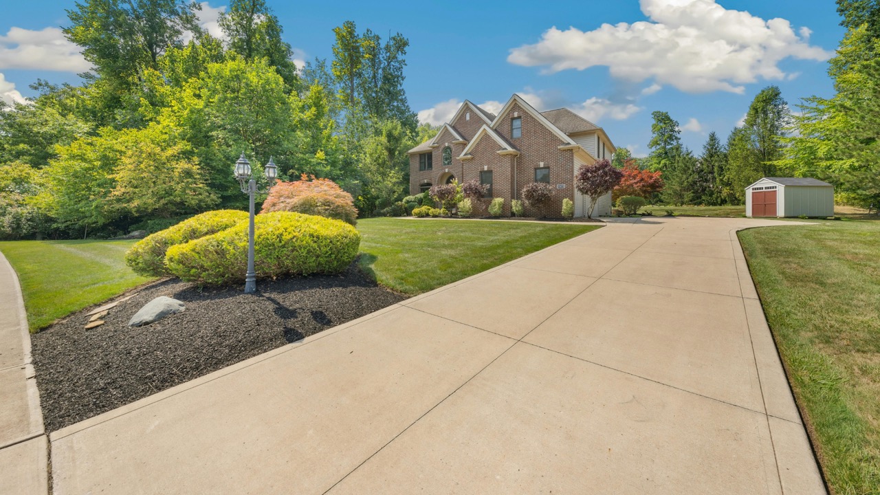 Spacious concrete driveway leading to brick home with manicured lawn, landscaped gardens, and detached garage on sunny day
