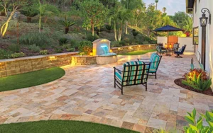 Luxury backyard patio with stone pavers, striped lounge chairs, illuminated retaining walls, and manicured landscape featuring tropical plants and palm trees