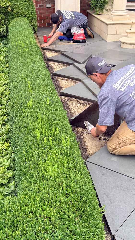 Two workers installing and sealing pavers along a residential walkway next to green boxwood hedges in Fond du Lac, Wisconsin