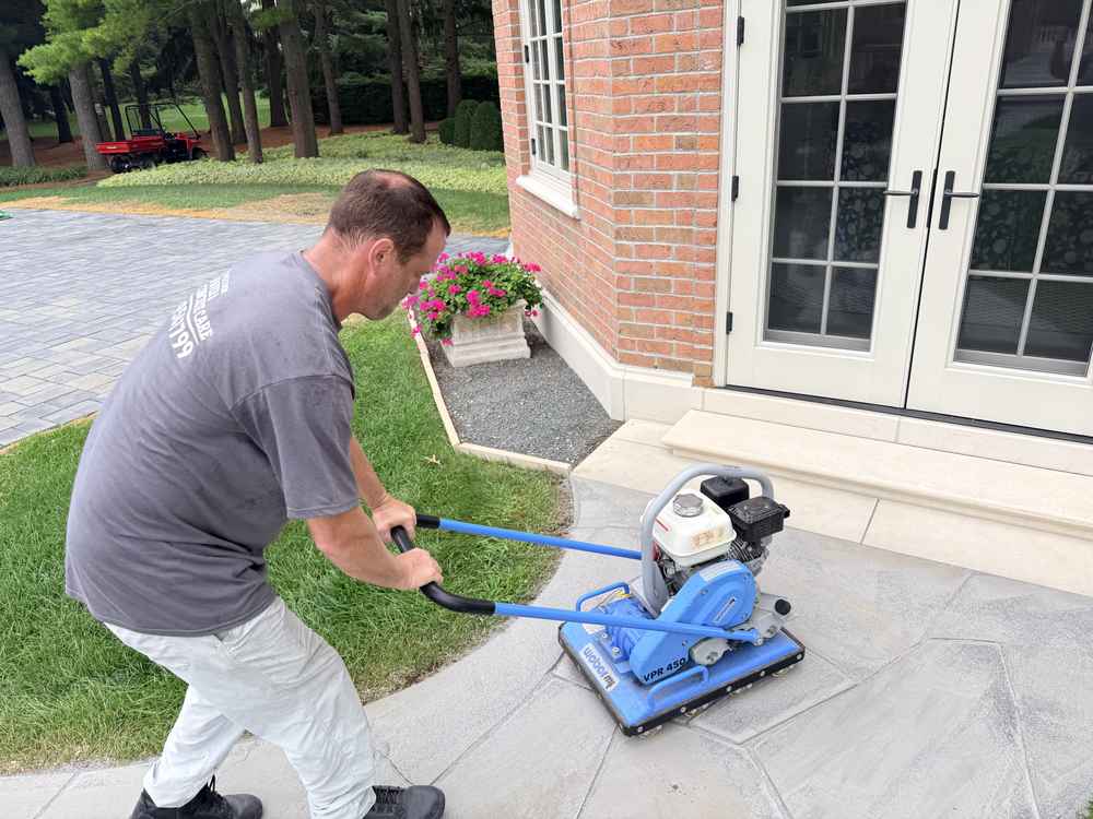 Man operating a blue paver sealing machine on a stamped concrete patio next to a brick home with flowering planters