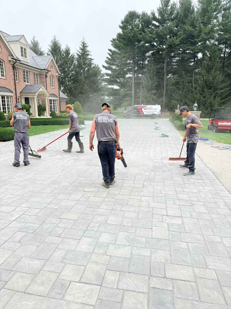 Four workers sealing and restoring a large paver driveway in front of a brick home in Fond du Lac, Wisconsin