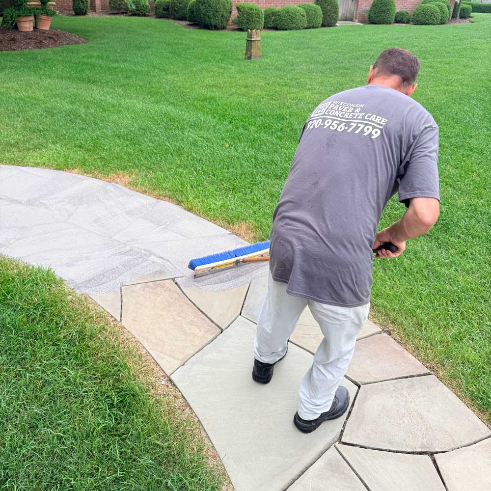 Worker cleaning concrete paver walkway with blue brush in Cincinnati residential yard with manicured lawn and hedges