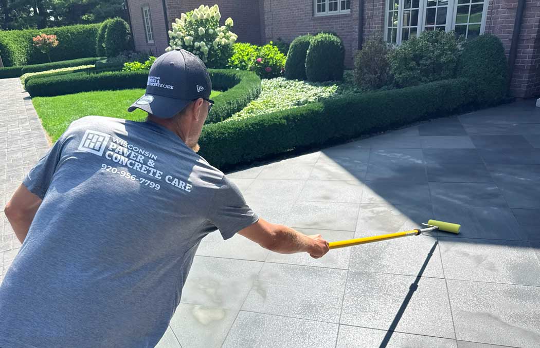 Worker in Wisconsin Concrete Care shirt cleaning driveway with long-handled brush tool, brick home and landscaped garden visible in background