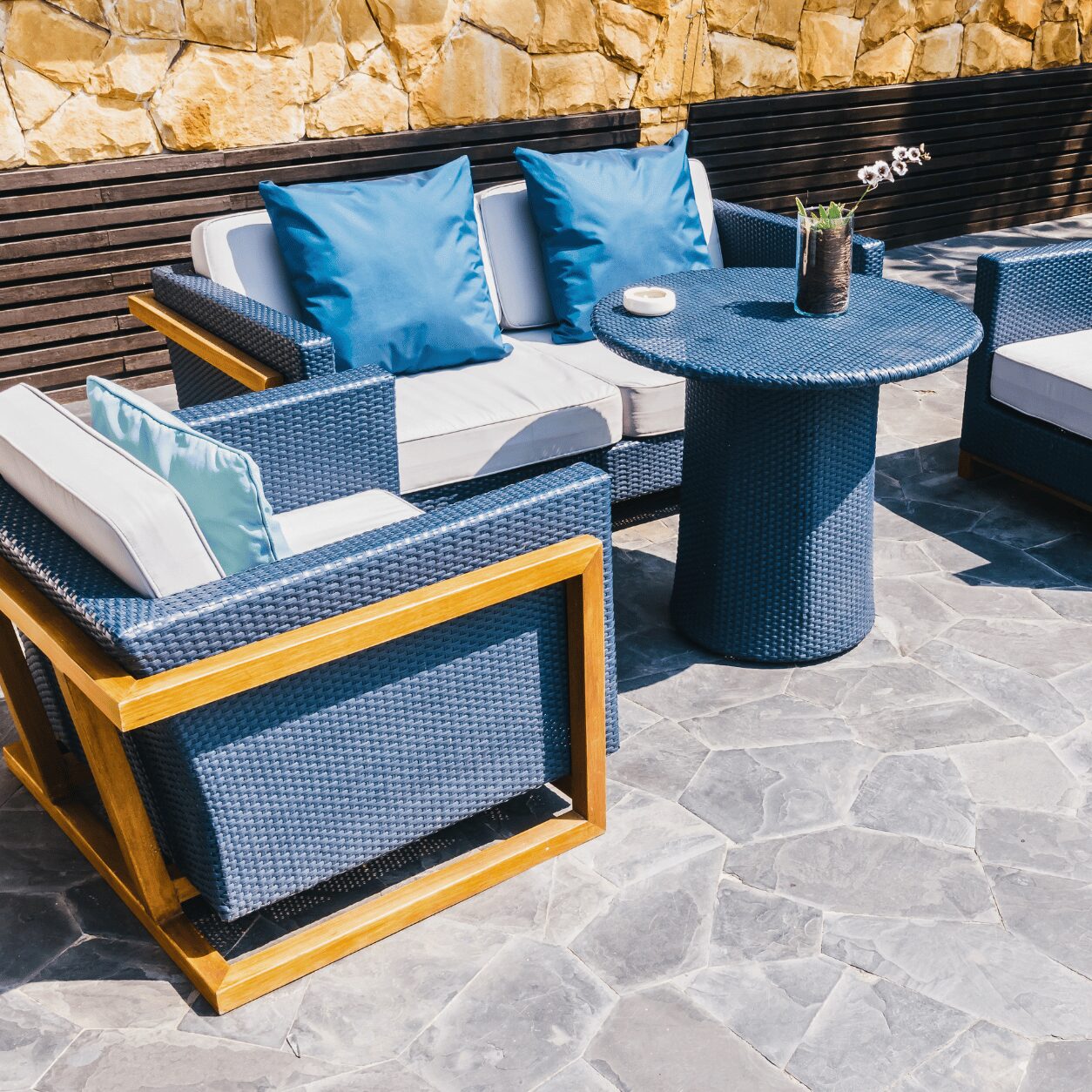 Modern outdoor patio seating area with blue wicker furniture and cushions on sealed pavers, featuring stone accent wall and wooden bench backdrop